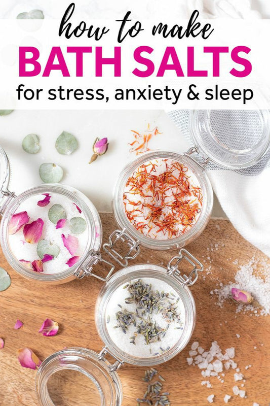 Three jars of bath salts with varying colors and inclusions, such as dried flowers and herbs, on a wooden surface with a text overlay that says 'how to make BATH SALTS for stress, anxiety & sleep'. The brand's website is mentioned as well.